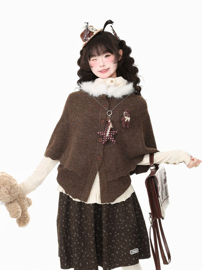 A model wearing a brown knit poncho cardigan with a white faux-fur collar, styled with a cream long-sleeve shirt, a dark brown patterned skirt, and a brown cat-themed headband, holding a brown teddy bear and a small brown bag against a plain white background.