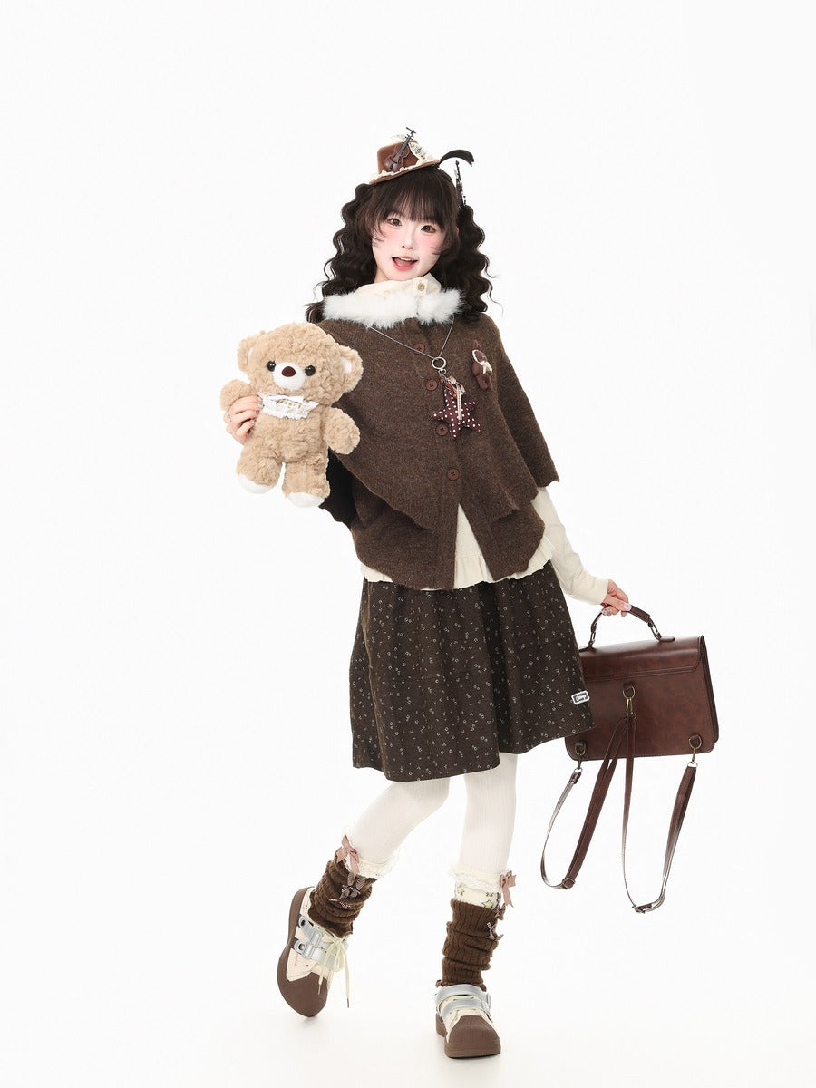 A model wearing a brown bohemian-style outfit with a patterned skirt, brown cardigan, and brown hat, holding a teddy bear and carrying a brown handbag against a white background.