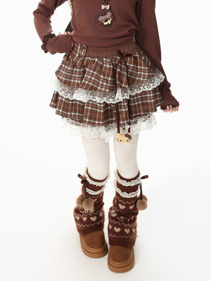 A child wearing a brown plaid tiered skirt with white lace trim, a matching brown sweater, white tights, and brown knit leg warmers with pom-poms and heart patterns, photographed against a white studio background.