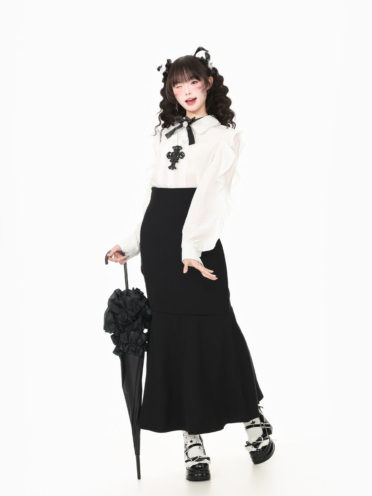 A model posing in a black and white outfit, featuring a long-sleeve white blouse with a black bow and cross accent, a black skirt, black platform shoes, and holding a black flower-covered umbrella against a plain white background.