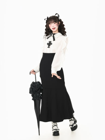 A model posing in a black and white outfit, featuring a long-sleeve white blouse with a black bow and cross accent, a black skirt, black platform shoes, and holding a black flower-covered umbrella against a plain white background.