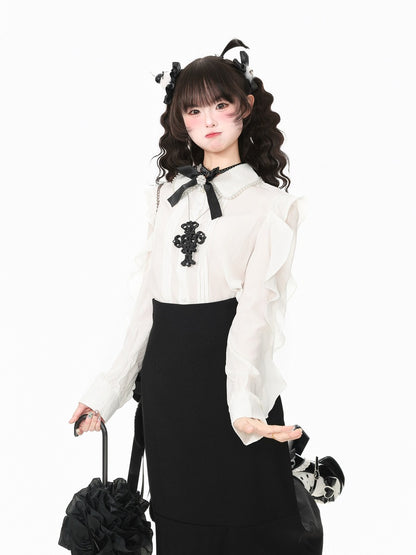 A young woman modeling a black and white gothic Lolita outfit, featuring a white long-sleeve blouse with a black bow and cross embellishment, a black skirt, and matching black hair accessories, set against a plain white background.