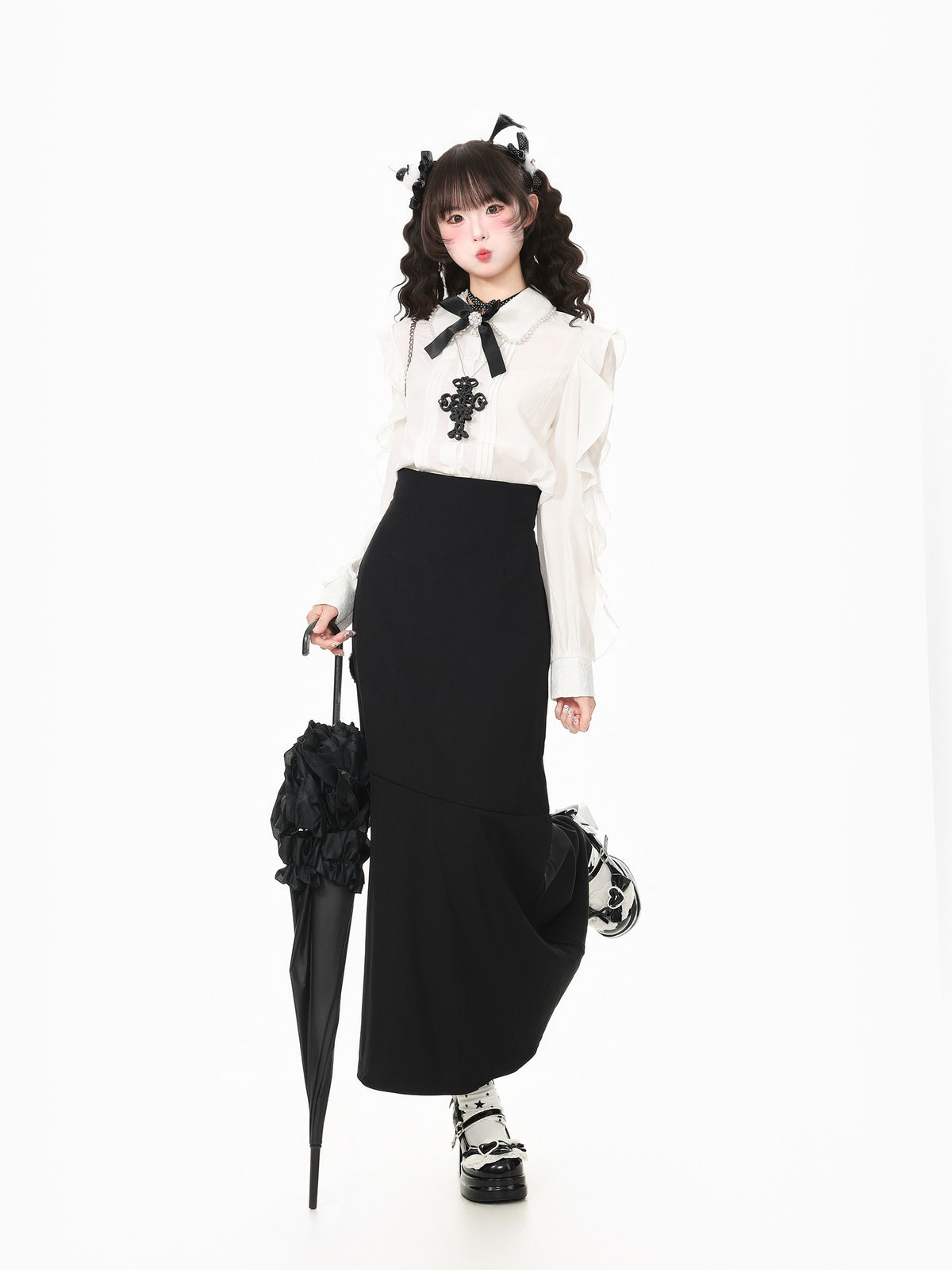 A model posing in a white long-sleeve blouse with a black bow and cross emblem, paired with a high-waisted black skirt, holding a black umbrella against a solid white background.