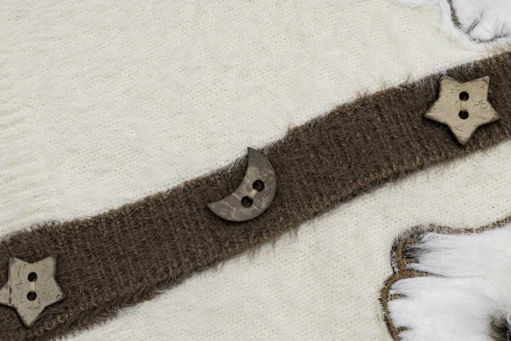 A close-up of a brown fuzzy knit trim or belt laid diagonally across a cream knit fabric, decorated with wooden star and crescent moon buttons.