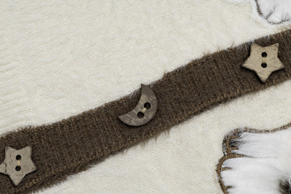 A close-up of a brown fuzzy knit trim or belt laid diagonally across a cream knit fabric, decorated with wooden star and crescent moon buttons.