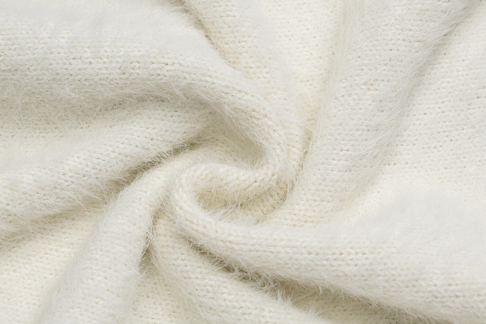 A close-up of soft, fluffy knit fabric in a warm off-white or cream color, arranged in a gentle swirl to show texture and drape.