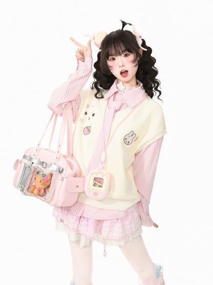 A young woman posing in a cute pastel pink and cream Lolita-style outfit, featuring a cream vest with cartoon patches, a pink gingham long-sleeve shirt, a pleated pink skirt, and carrying a pink bag with a clear front window.