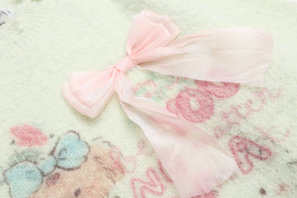 A light pink sheer organza bow placed on a soft, pale green blanket with a cartoon bear and pink letter pattern.
