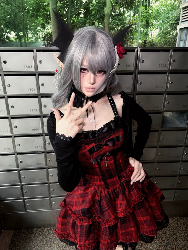 A person wearing a gothic Lolita-style dress with a red and black plaid skirt, black long sleeves, and a black lace collar, posing in front of a row of gray mailboxes outdoors.