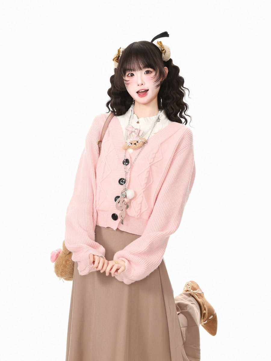 A young woman models a cute pink ribbed cardigan sweater with black buttons, styled with a brown pleated skirt, a white collar shirt, and a teddy bear accessory, set against a plain white background.