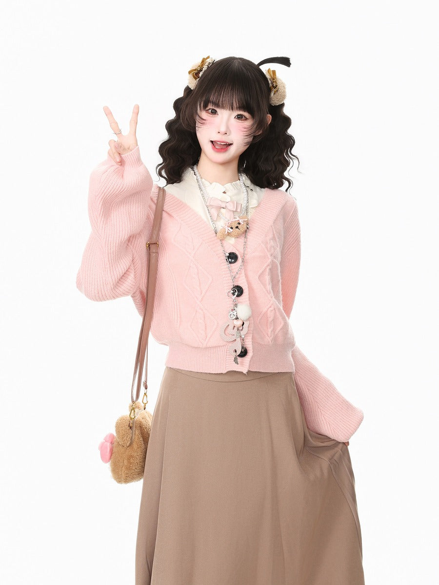 A young woman posing in a light pink cable-knit cardigan with white trim and decorative buttons, paired with a beige maxi skirt. She is accessorized with a small pink and brown plush shoulder bag, cat ears headband, and cat-eye makeup against a solid white background.