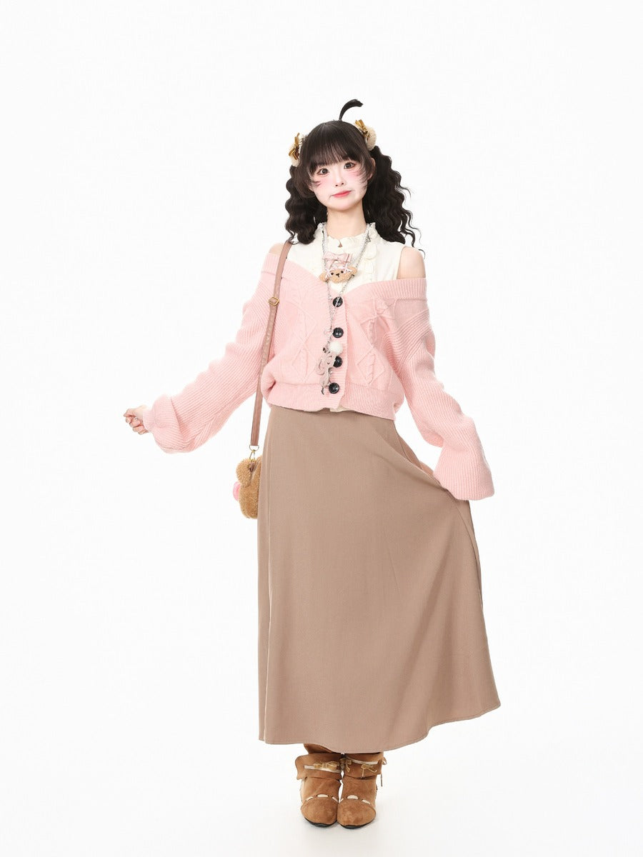 A model wearing a light pink cropped cardigan with black buttons, a beige maxi skirt, a white collar blouse, and brown boots, set against a plain white background.