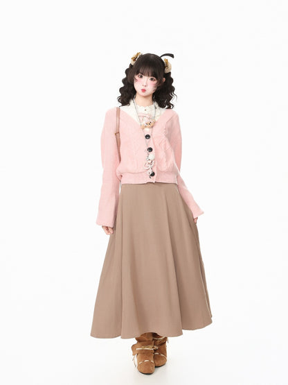 A model wearing a light pink cropped cardigan with black buttons, a beige maxi skirt, and brown shoes, standing against a plain white background.