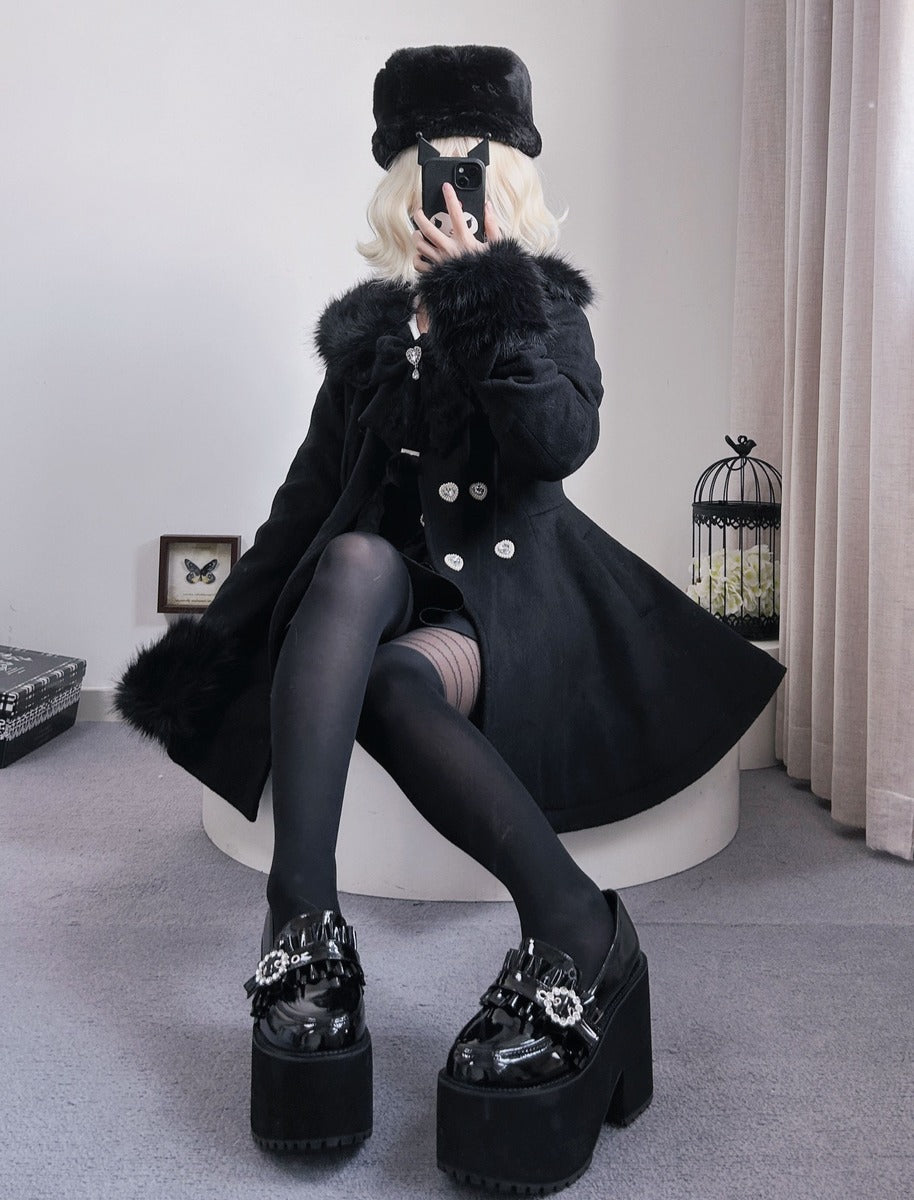 A person wearing a black fur-trimmed coat, black hat, black tights, and black platform shoes with rhinestone embellishments, seated on a white stool in an indoor room.