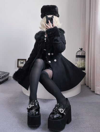 A person wearing a black fur-trimmed coat, black hat, black tights, and black platform shoes with rhinestone embellishments, seated on a white stool in an indoor room.