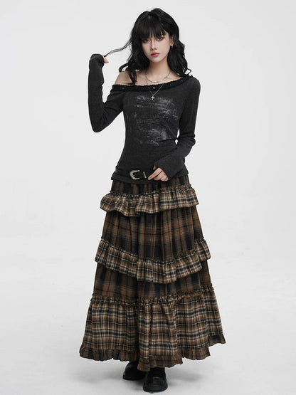 A model wearing a gothic-style outfit consisting of a black off-the-shoulder long-sleeve top and a long brown and black plaid tiered ruffle skirt, standing against a plain white studio background.