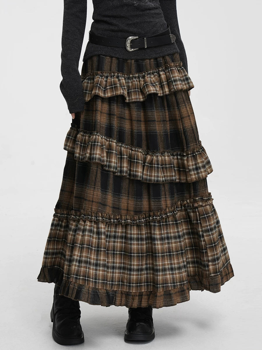 A long tiered ruffle skirt in a brown, black, and beige plaid pattern, styled with a black belt, dark gray long-sleeve top, and black boots against a light gray studio background.