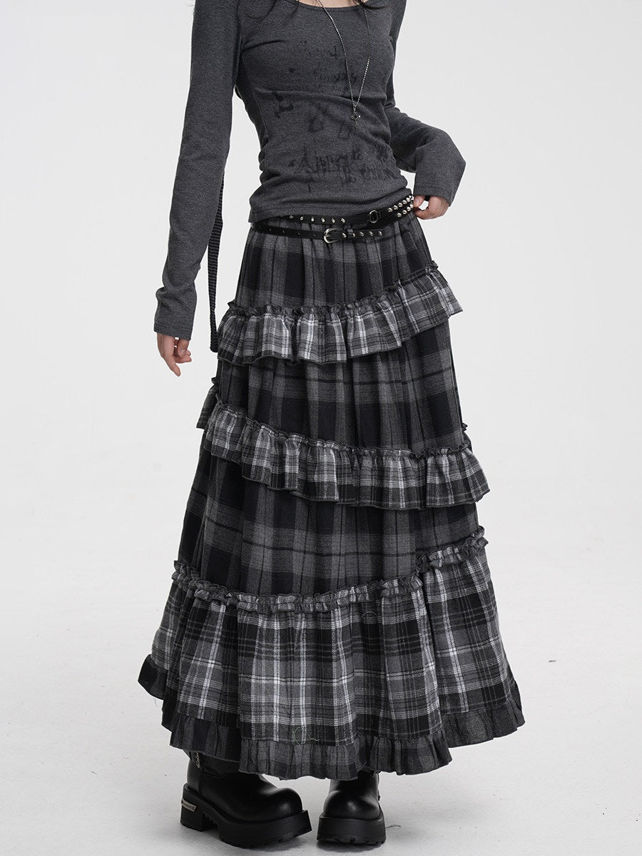 A model wearing a long, tiered black and gray plaid skirt with ruffles, paired with a dark gray long-sleeve top and black platform boots, against a plain white studio background.