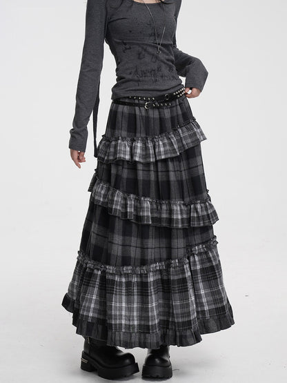 A model wearing a long, tiered black and gray plaid skirt with ruffles, paired with a dark gray long-sleeve top and black platform boots, against a plain white studio background.