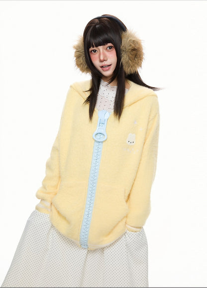 A young woman wearing a fuzzy pale yellow hooded jacket with a light blue zipper, tan faux-fur ear muffs, and a white polka-dot skirt against a plain white background.