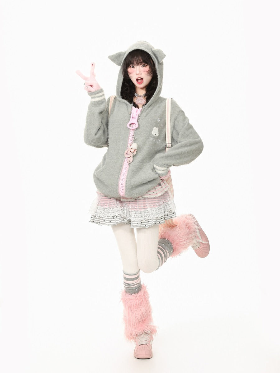 A model posing in a light gray cat-ear hooded sweatshirt with a pink zipper and cute accessories, paired with a patterned pleated skirt, pink fuzzy socks, and pink boots, set against a plain white background.