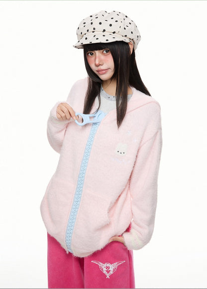 A model wearing a fuzzy light pink zip-up hoodie with a light blue trim and a small white bear face on the chest, paired with pink pants and a white polka-dot cap, against a plain white background.