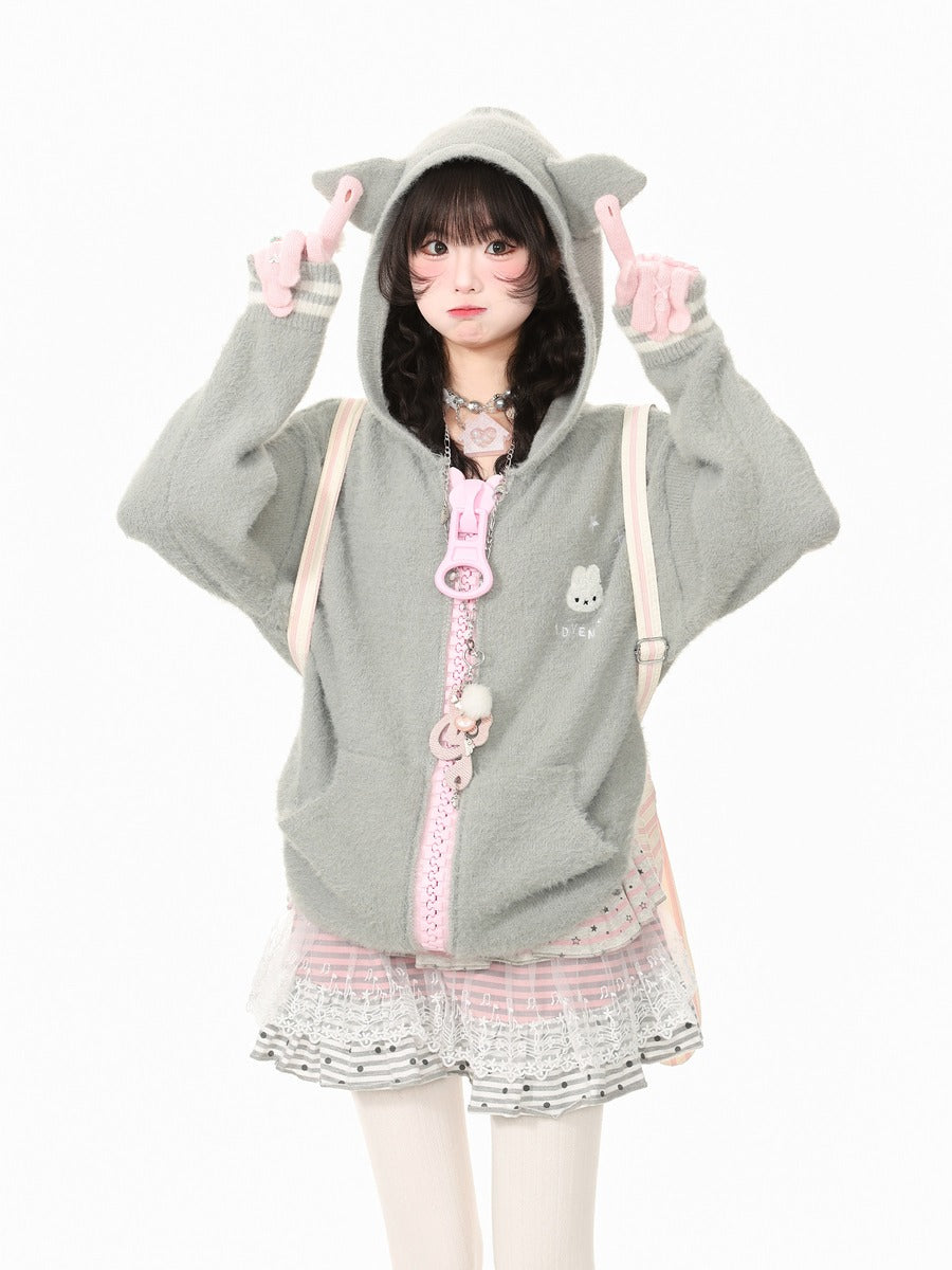 A young woman modeling a light gray fuzzy hooded sweatshirt with pink accents, including a pink zipper, a pink bunny charm, and a pink-and-white striped pleated skirt, posing against a solid white background.