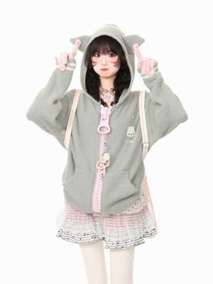 A young woman modeling a light gray fuzzy hooded sweatshirt with pink accents, including a pink zipper, a pink bunny charm, and a pink-and-white striped pleated skirt, posing against a solid white background.