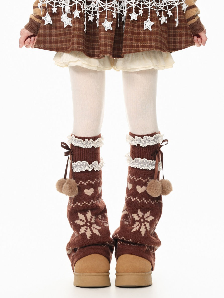 A person shown from the waist down wearing brown knit leg warmers with cream snowflake and heart patterns, white lace trim, and pom-poms, paired with white tights and a brown plaid skirt.