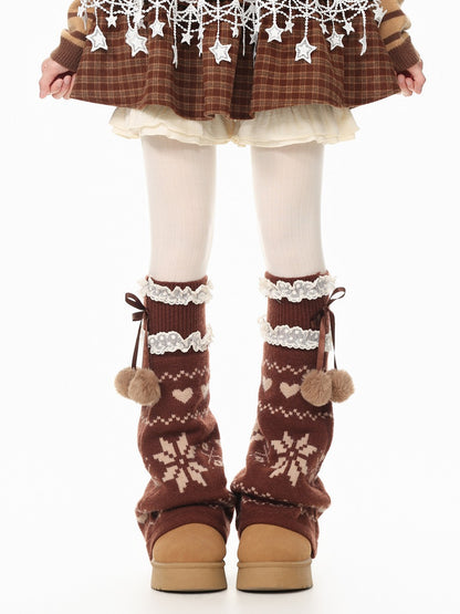 A person shown from the waist down wearing brown knit leg warmers with cream snowflake and heart patterns, white lace trim, and pom-poms, paired with white tights and a brown plaid skirt.