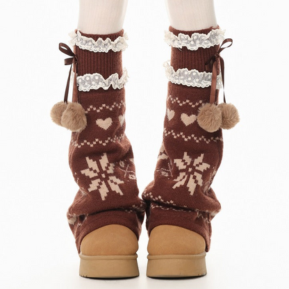 Brown knit leg warmers with cream snowflake and heart patterns, white lace trim, and pom-pom details, shown on a white background.
