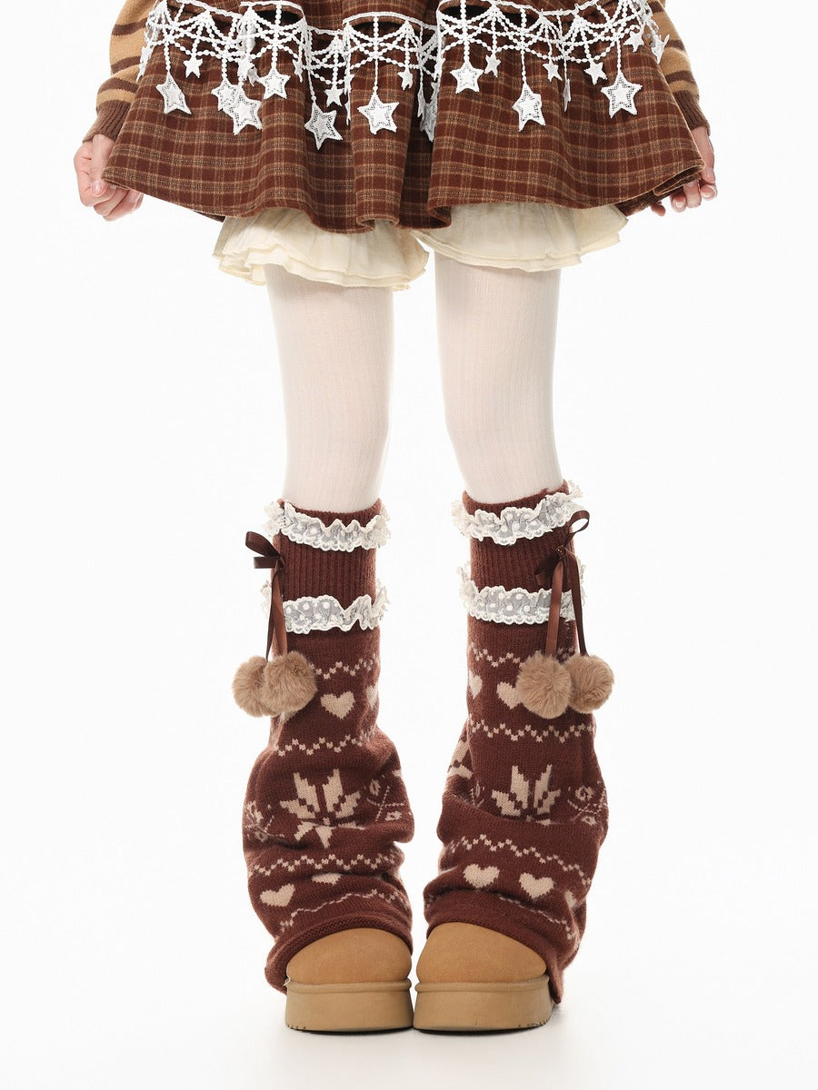 A child's legs wearing brown knit leg warmers with cream patterns, lace trim, and pom-poms, paired with brown boots, white tights, and a brown plaid skirt with white star details.