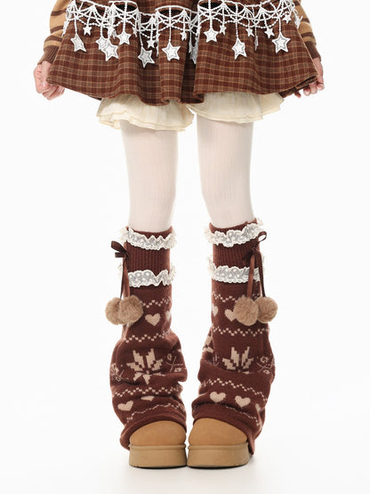A child's legs wearing brown knit leg warmers with cream patterns, lace trim, and pom-poms, paired with brown boots, white tights, and a brown plaid skirt with white star details.