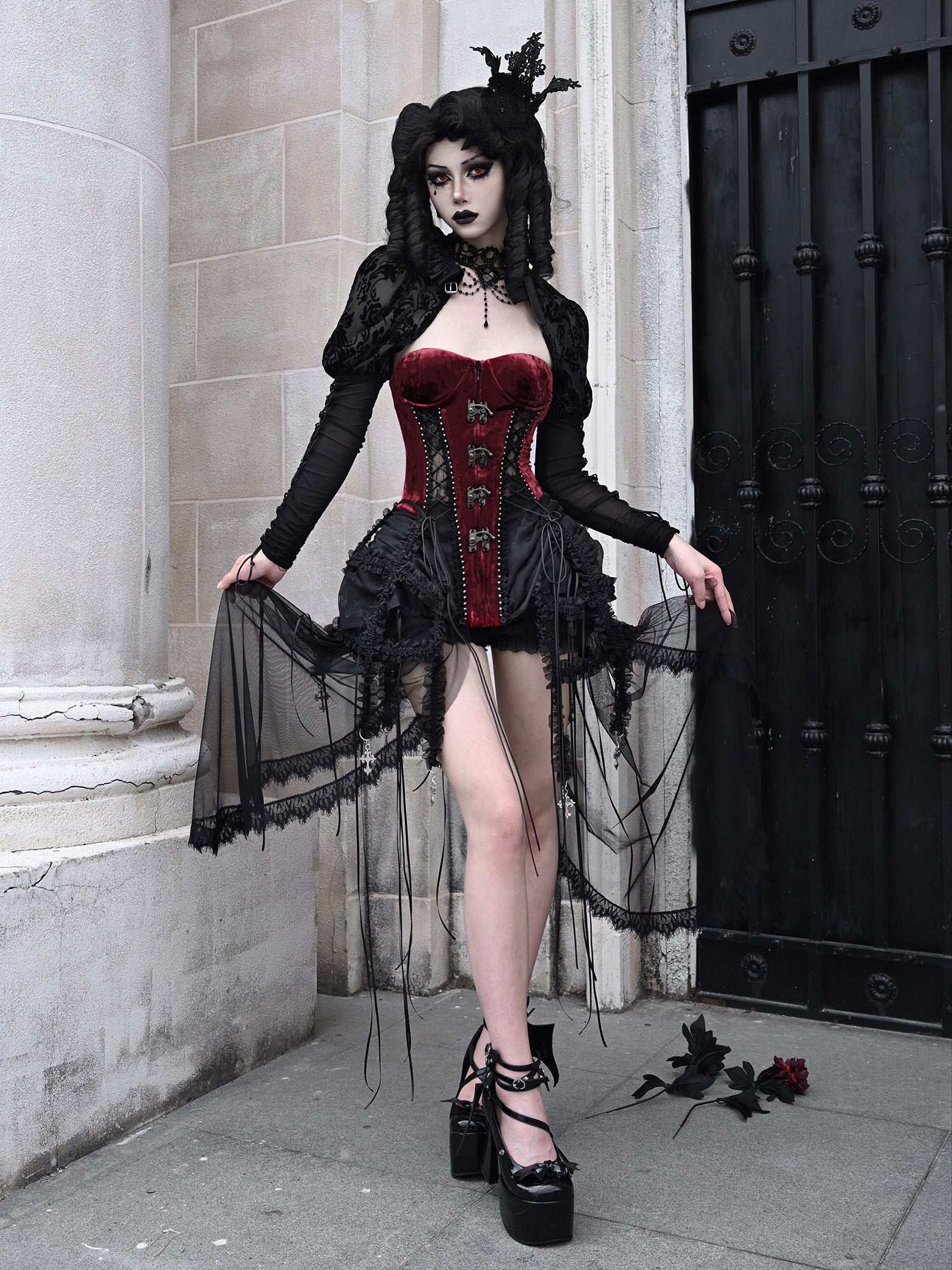 Person wearing a gothic-style outfit with a red corset and black lace dress, standing against a stone wall.