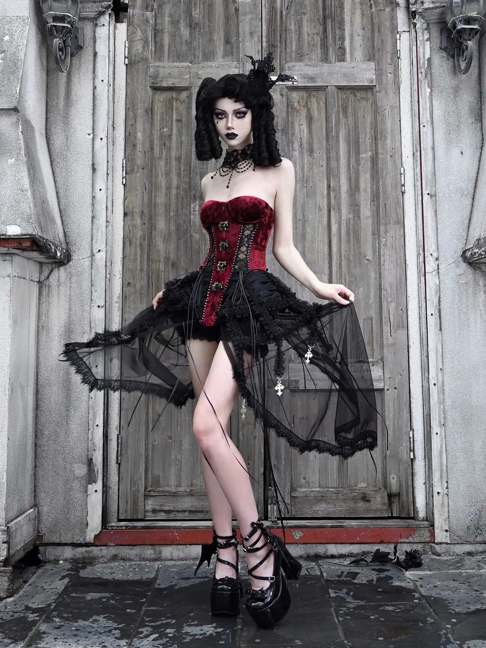 Woman in a gothic outfit with a red corset and black skirt standing in front of a wooden door.