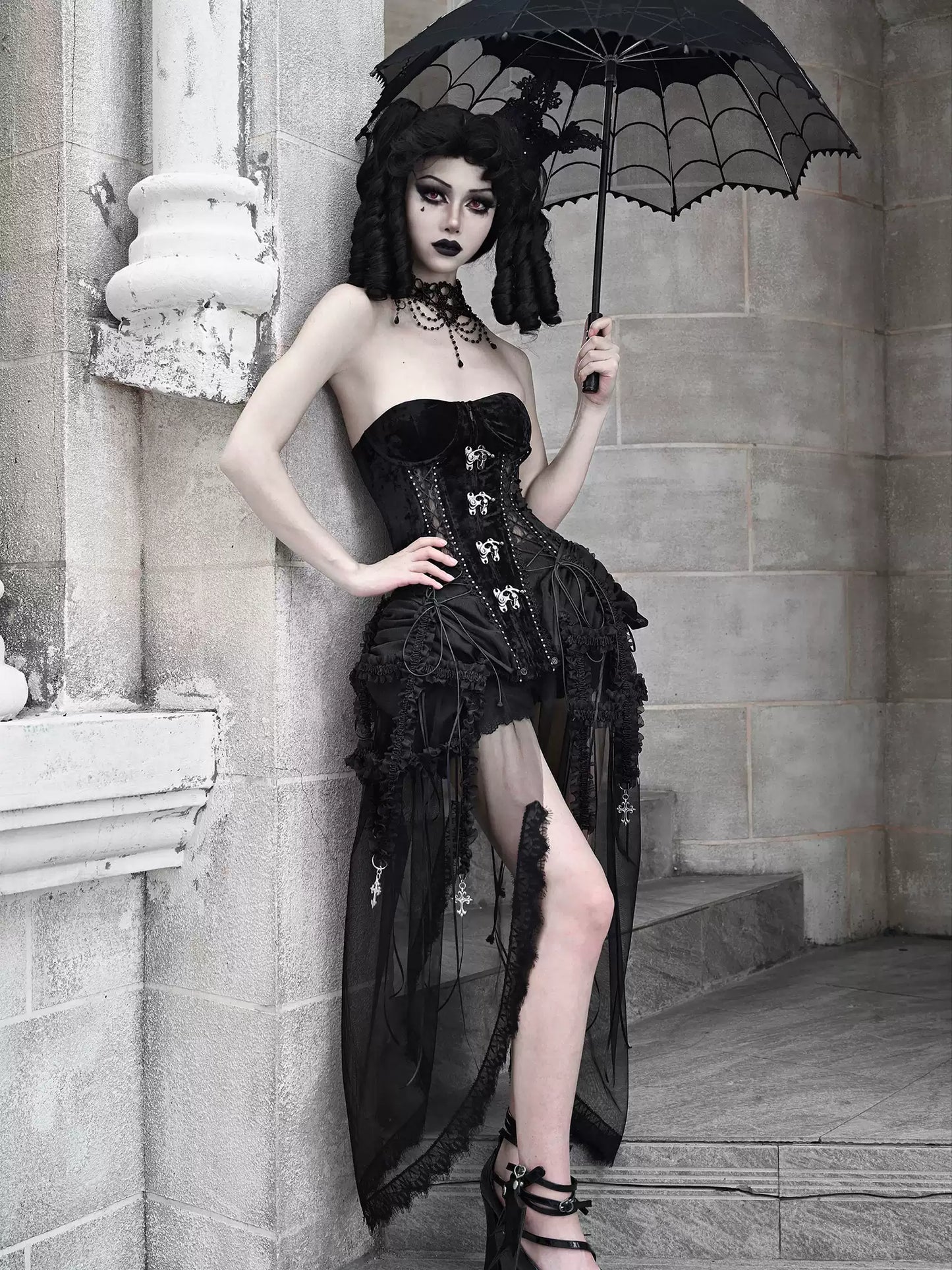 Gothic woman in a black corset and skirt holding a black lace umbrella against a stone wall.