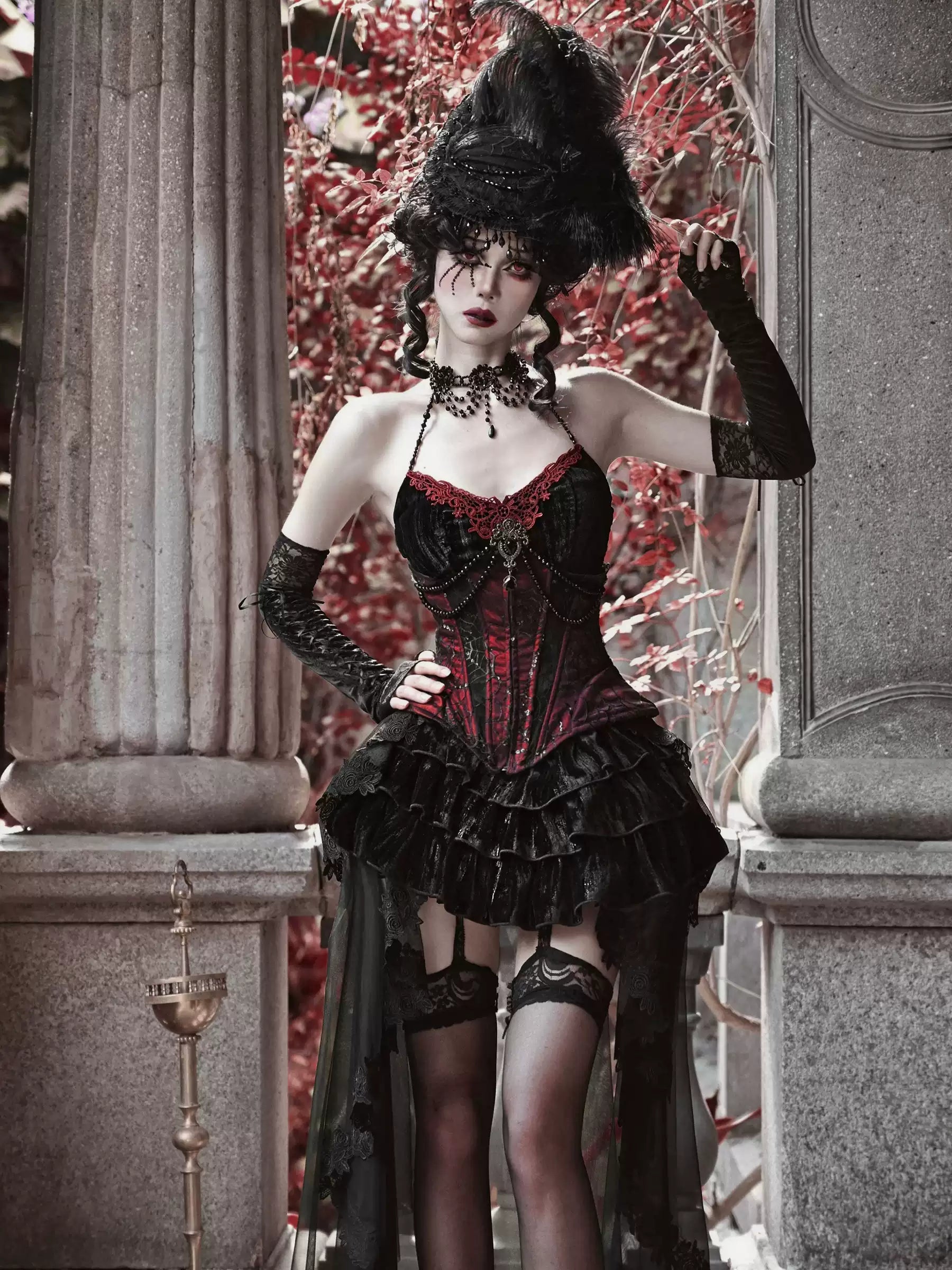 Woman in a gothic-style outfit with a black hat and red corset standing in an ornate setting.