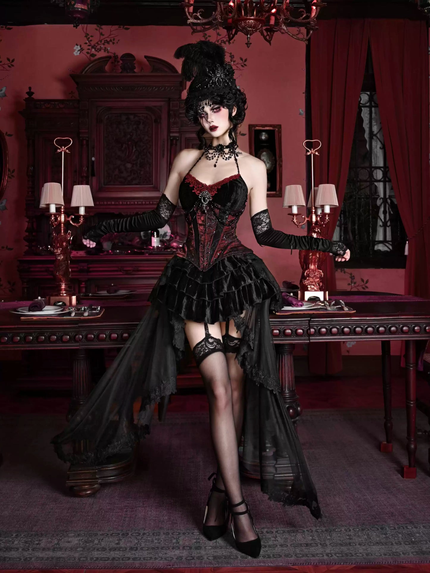 Woman in a black Victorian-style dress standing in a dimly lit room with red walls and a chandelier.