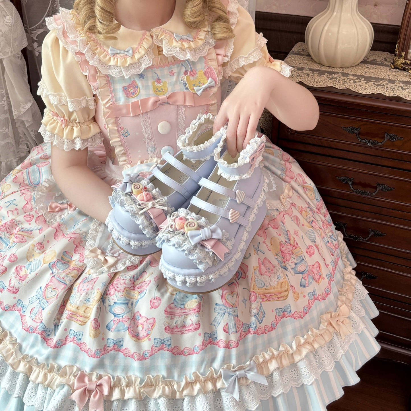 Child in a detailed dress holding blue lolita shoes with a wooden dresser in the background