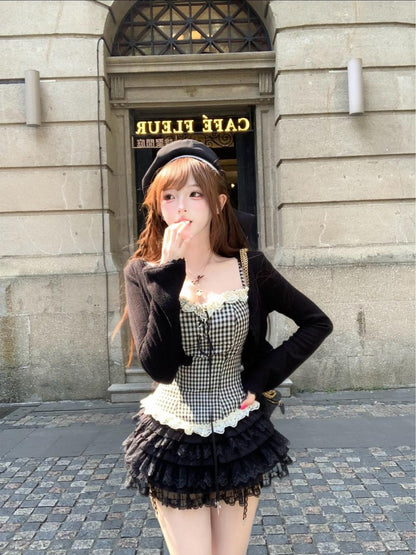 Woman in a dark coquette black outfit with a checkered apron standing in front of a building entrance.