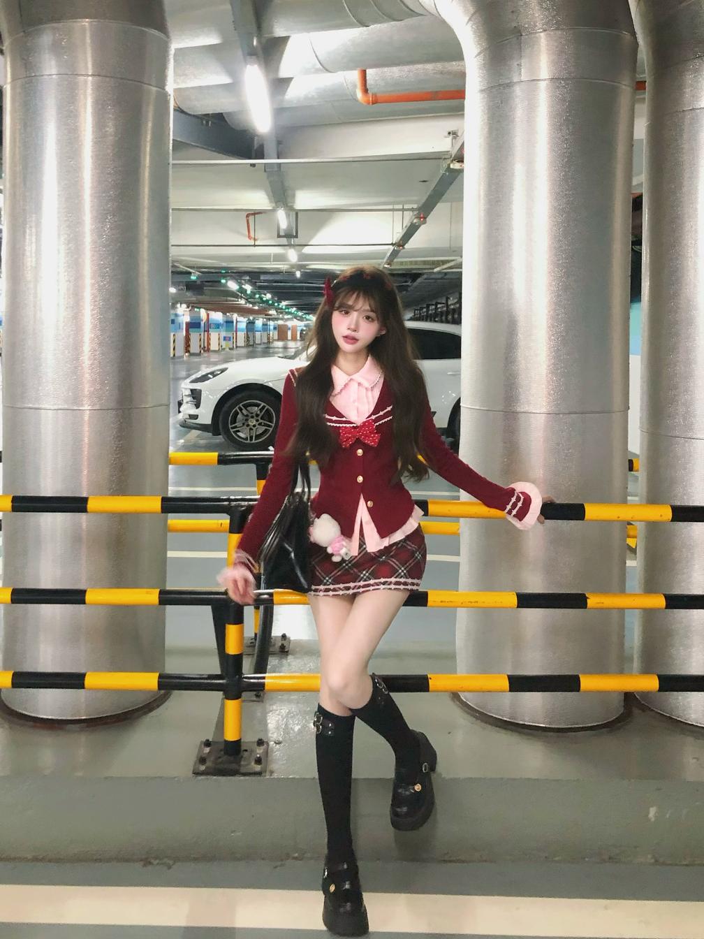 Person in a red school uniform standing in an indoor setting with large cylindrical columns and a vehicle in the background.