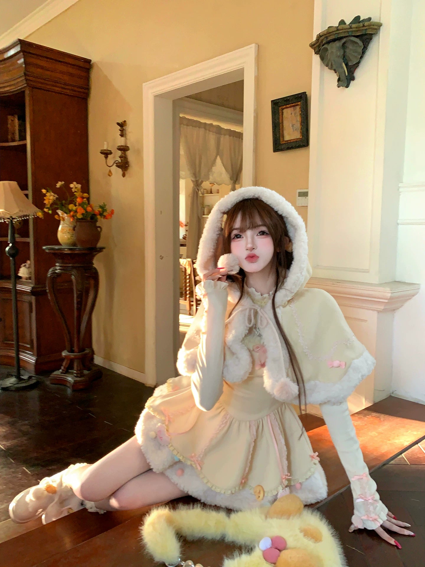 Person in a cream-colored outfit with a hood sitting on a wooden floor in a room.
