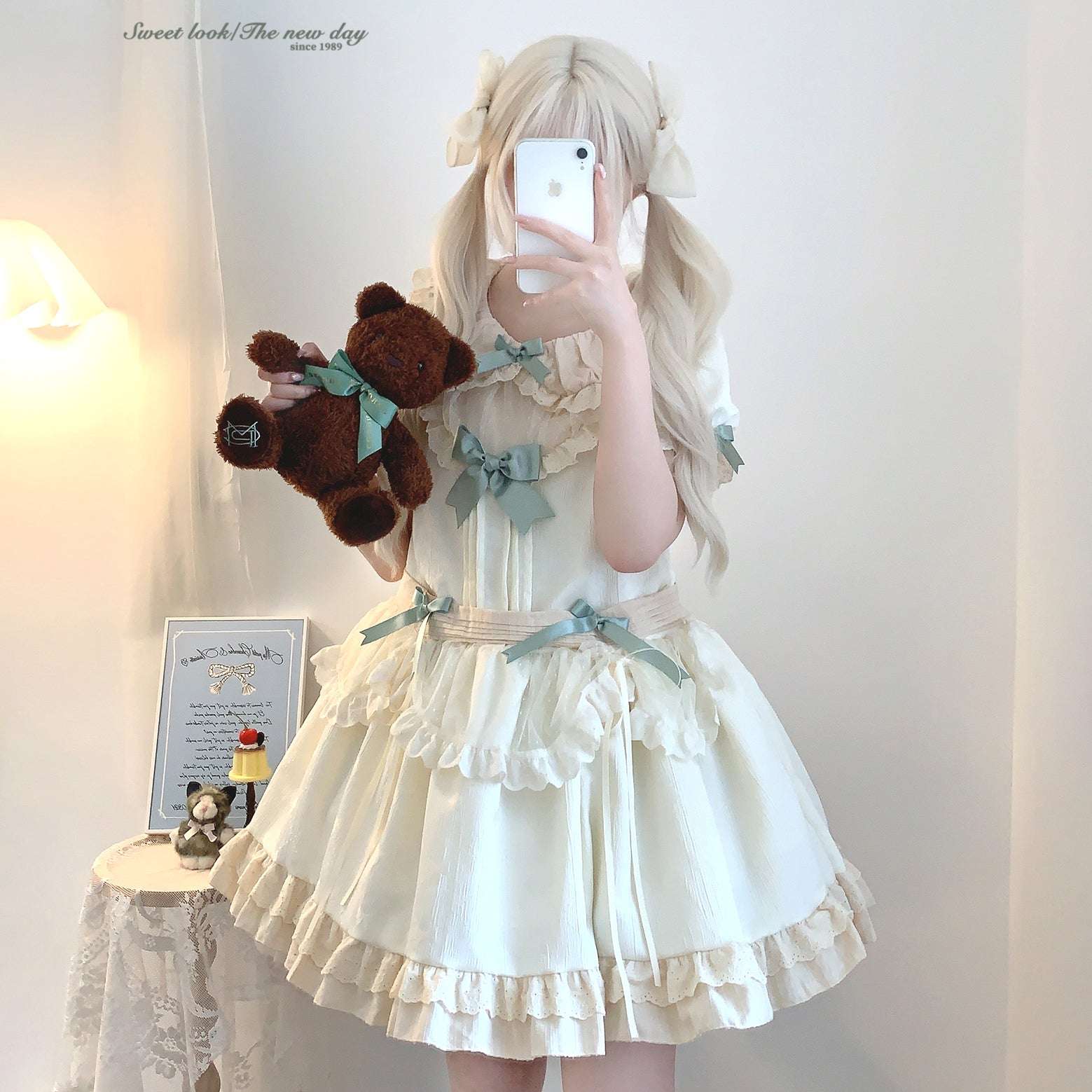 Person wearing a white dress with ruffles and bows, holding a teddy bear, taking a mirror selfie.