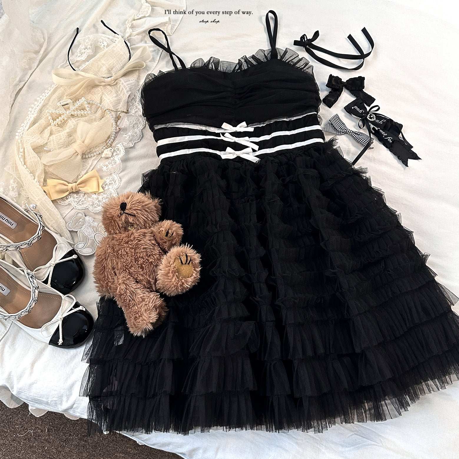 Black ruffled dress with accessories including a teddy bear, shoes, and ribbons on a white surface.