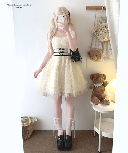 Person wearing a white dress with black ribbons, standing in a room with a teddy bear and bags.