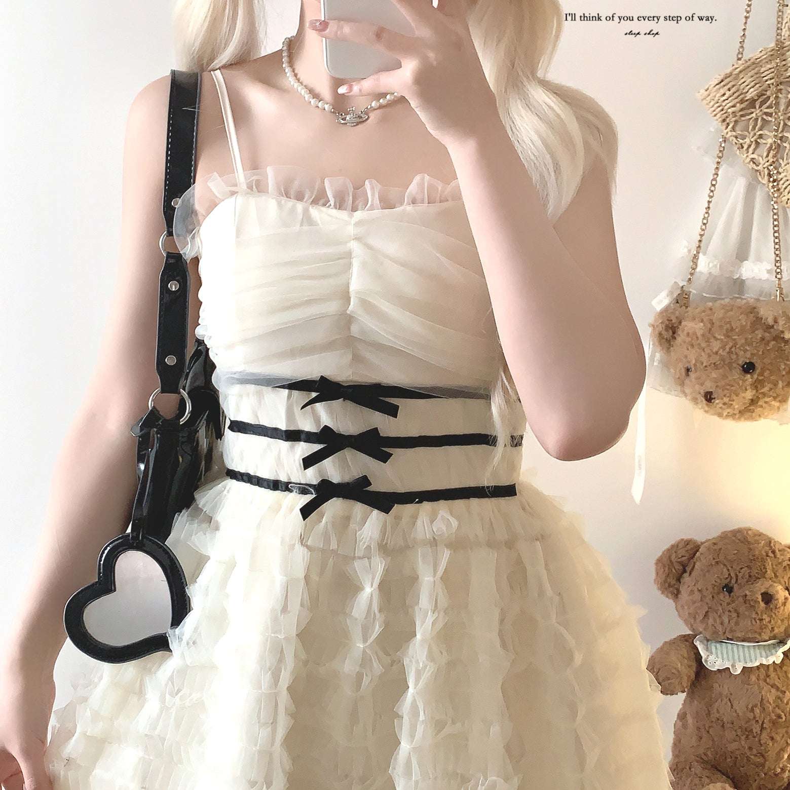 Person wearing a white dress with black ribbons and a black bag, standing next to teddy bears.