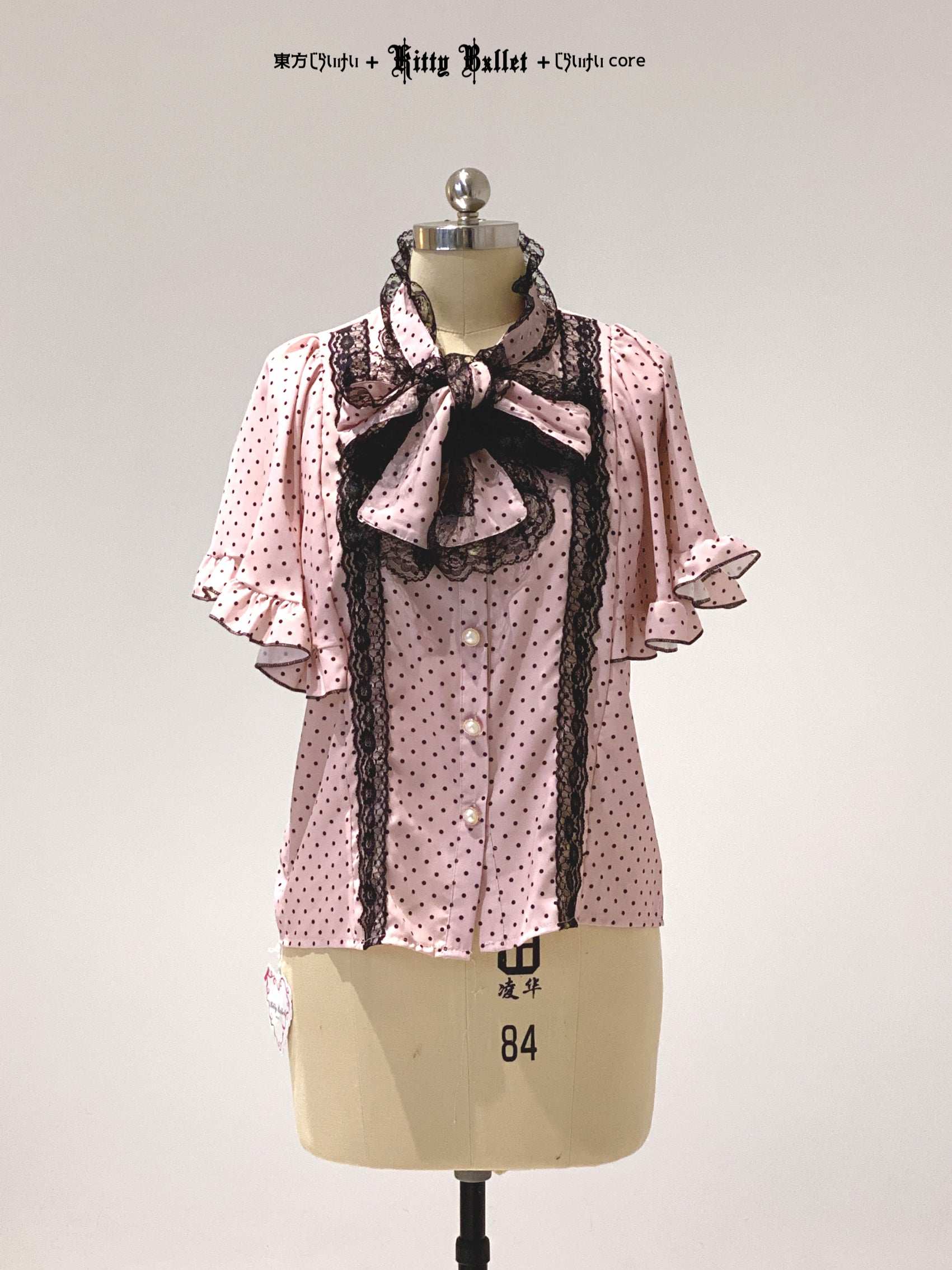 Pink polka dot blouse with black lace details on a mannequin against a gray background.
