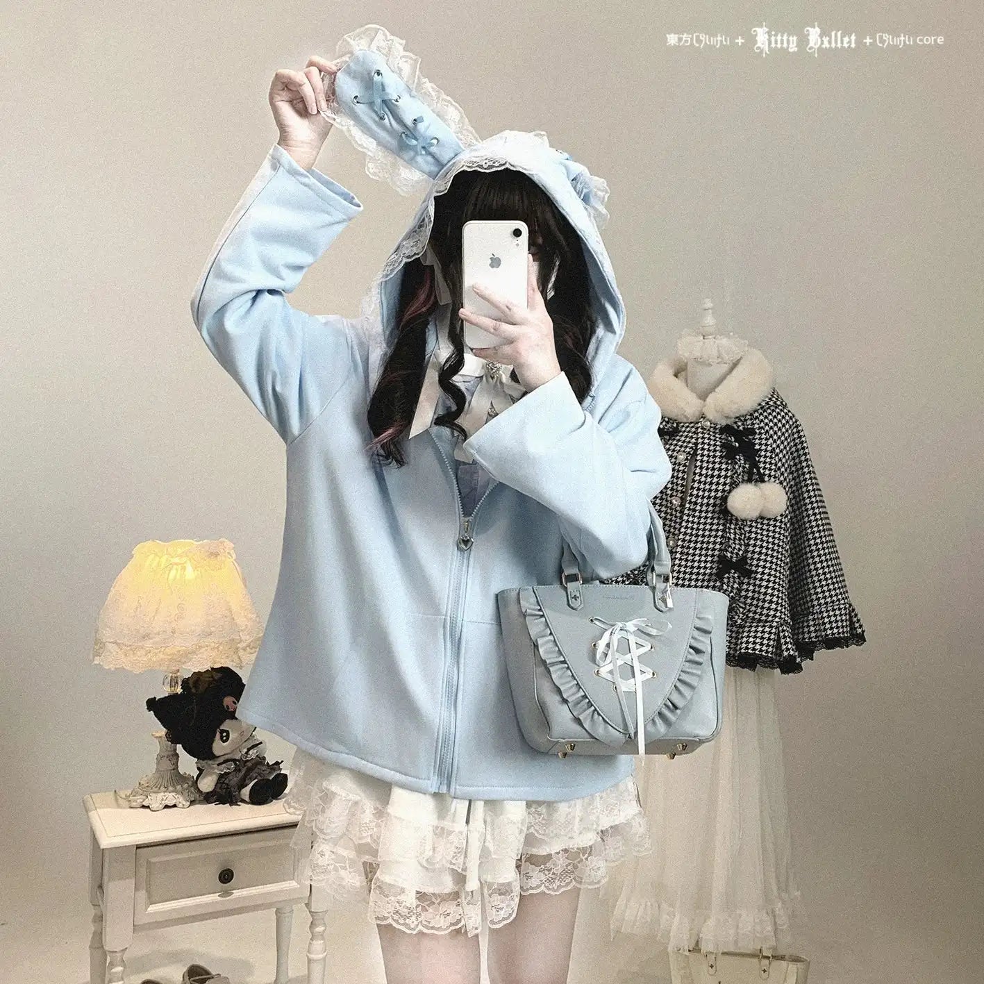 Person wearing a light blue hooded coat with ruffled details, holding a phone, in a room with a lamp and another outfit on a mannequin.