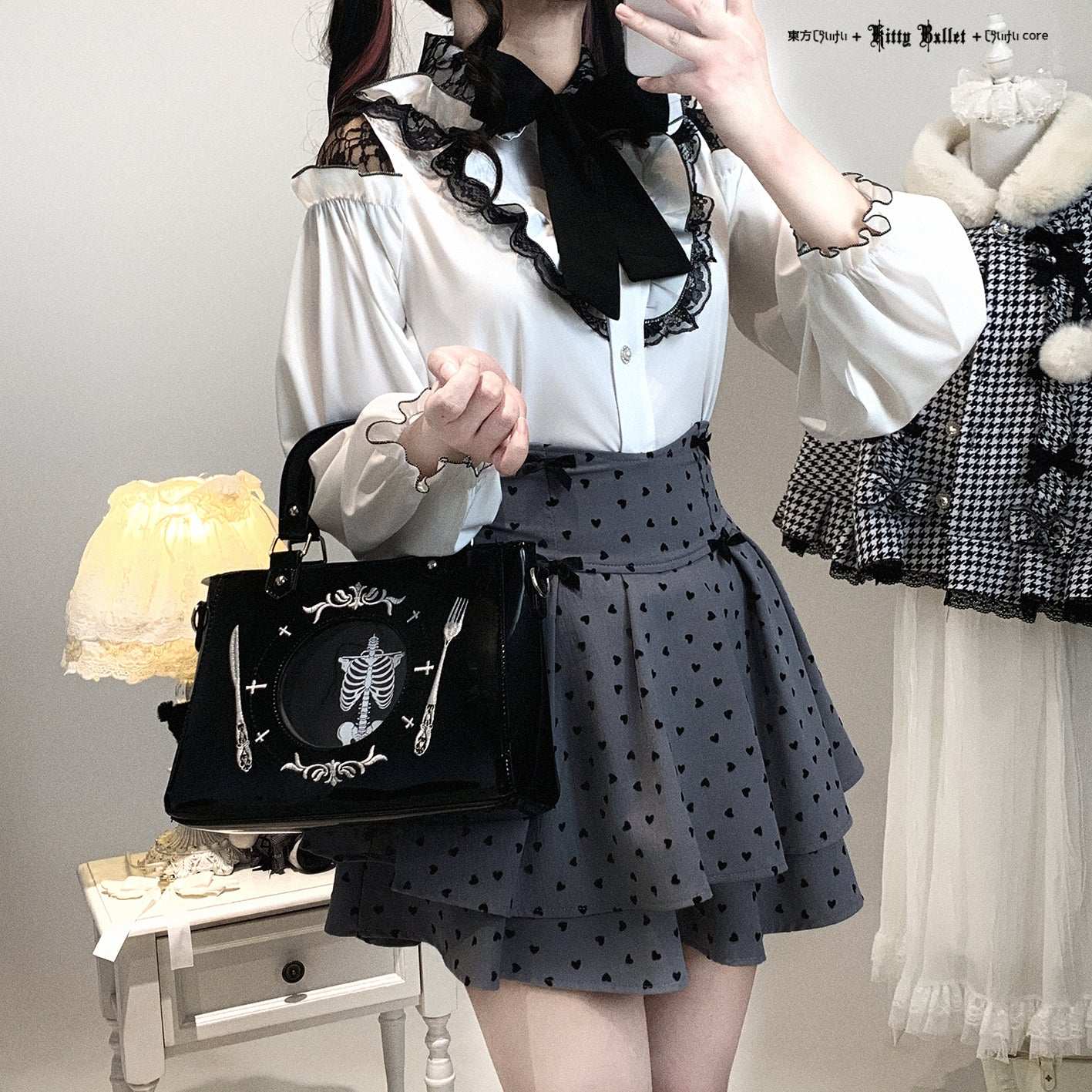 Person wearing a white blouse with black lace details and a polka dot skirt, holding a black bag with skeleton design.