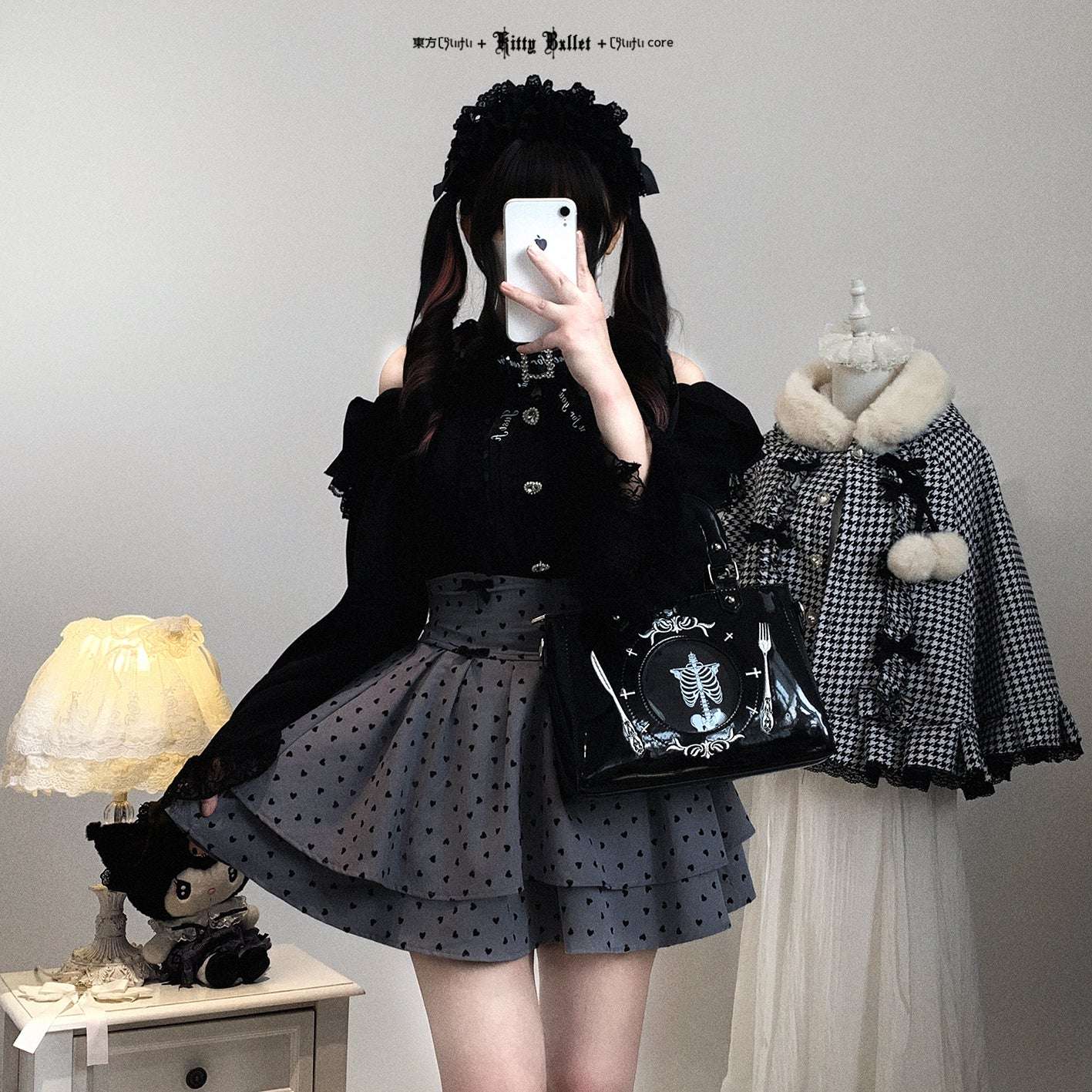 Person wearing a black and gray outfit with a polka dot skirt, holding a phone, in a room with a lamp and another outfit on a mannequin.